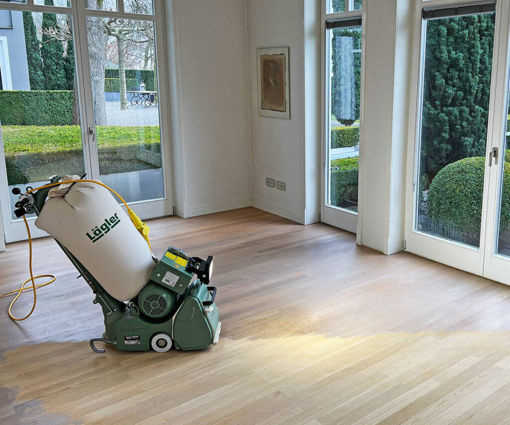 Bandschleifmaschine auf teilweise geschliffenem Parkett in einem hellen Wohnraum